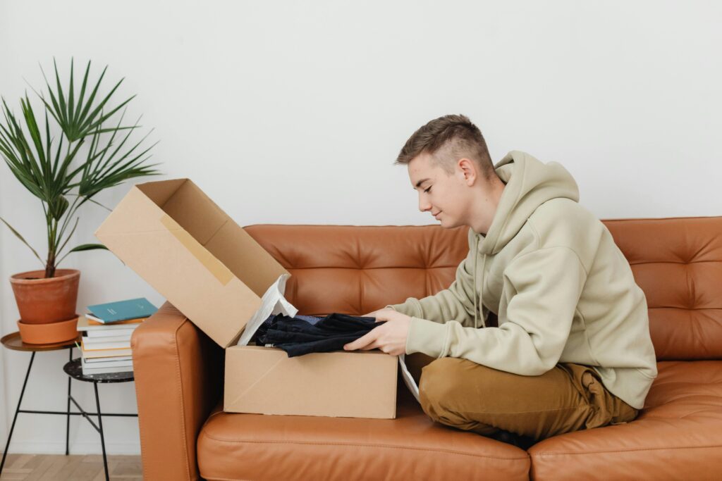 Young man in a hoodie unboxing a package while sitting on a brown leather sofa, surrounded by books and a potted plant.